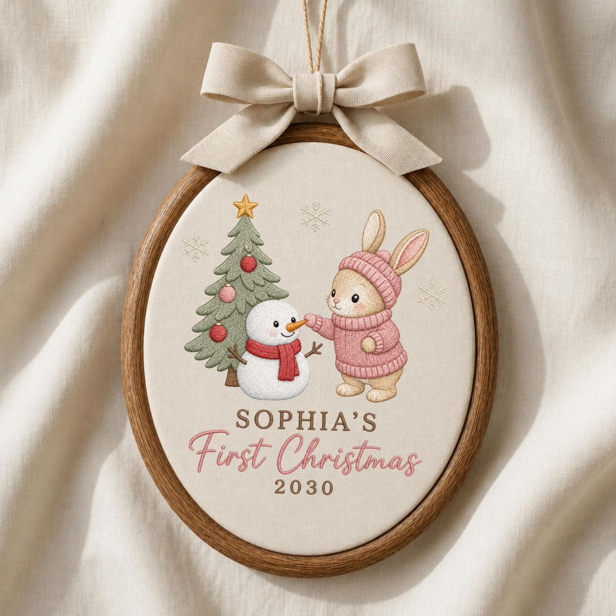 May include: An oval wooden embroidery hoop ornament featuring a beige fabric center with a printed illustration of a rabbit in a pink sweater, a snowman with a red scarf, and a Christmas tree. Text reads SOPHIA'S First Christmas 2030. A beige bow hangs above.