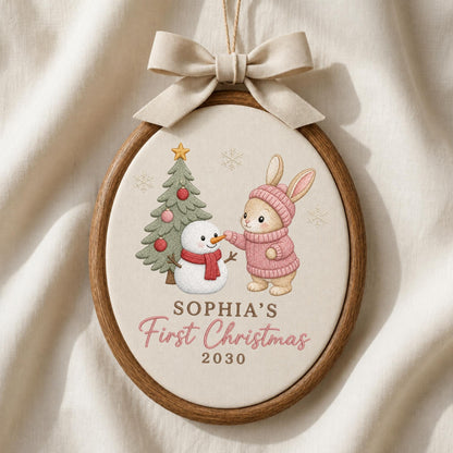 May include: An oval wooden embroidery hoop ornament featuring a beige fabric center with a printed illustration of a rabbit in a pink sweater, a snowman with a red scarf, and a Christmas tree. Text reads SOPHIA'S First Christmas 2030. A beige bow hangs above.