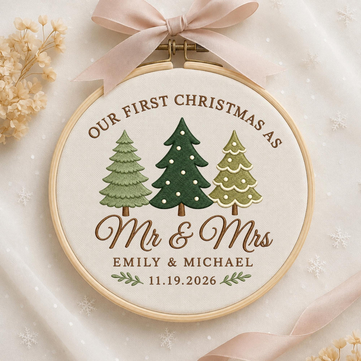 May include: A round wooden embroidery hoop containing cream fabric with the embroidered text "OUR FIRST CHRISTMAS AS Mr &amp; Mrs EMILY &amp; MICHAEL 11.19.2026" and three stylized green Christmas trees, topped with a light pink satin ribbon bow.