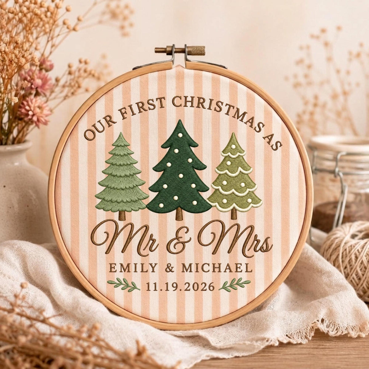 May include: A circular wooden embroidery hoop containing fabric with vertical beige stripes. The embroidery features three green pine trees, the text "OUR FIRST CHRISTMAS AS Mr &amp; Mrs", "EMILY &amp; MICHAEL", and the date "11.19.2026" with leaf accents.