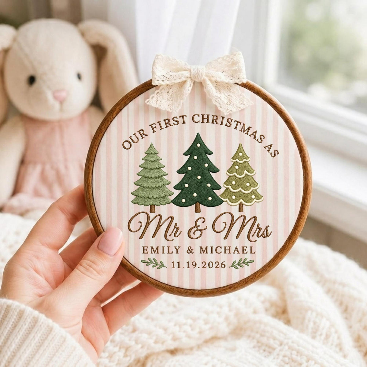 May include: A round wooden embroidery hoop ornament featuring a light pink and white striped fabric background. The design includes three embroidered pine trees, the text "OUR FIRST CHRISTMAS AS Mr &amp; Mrs EMILY &amp; MICHAEL 11.19.2026", and a cream lace bow.