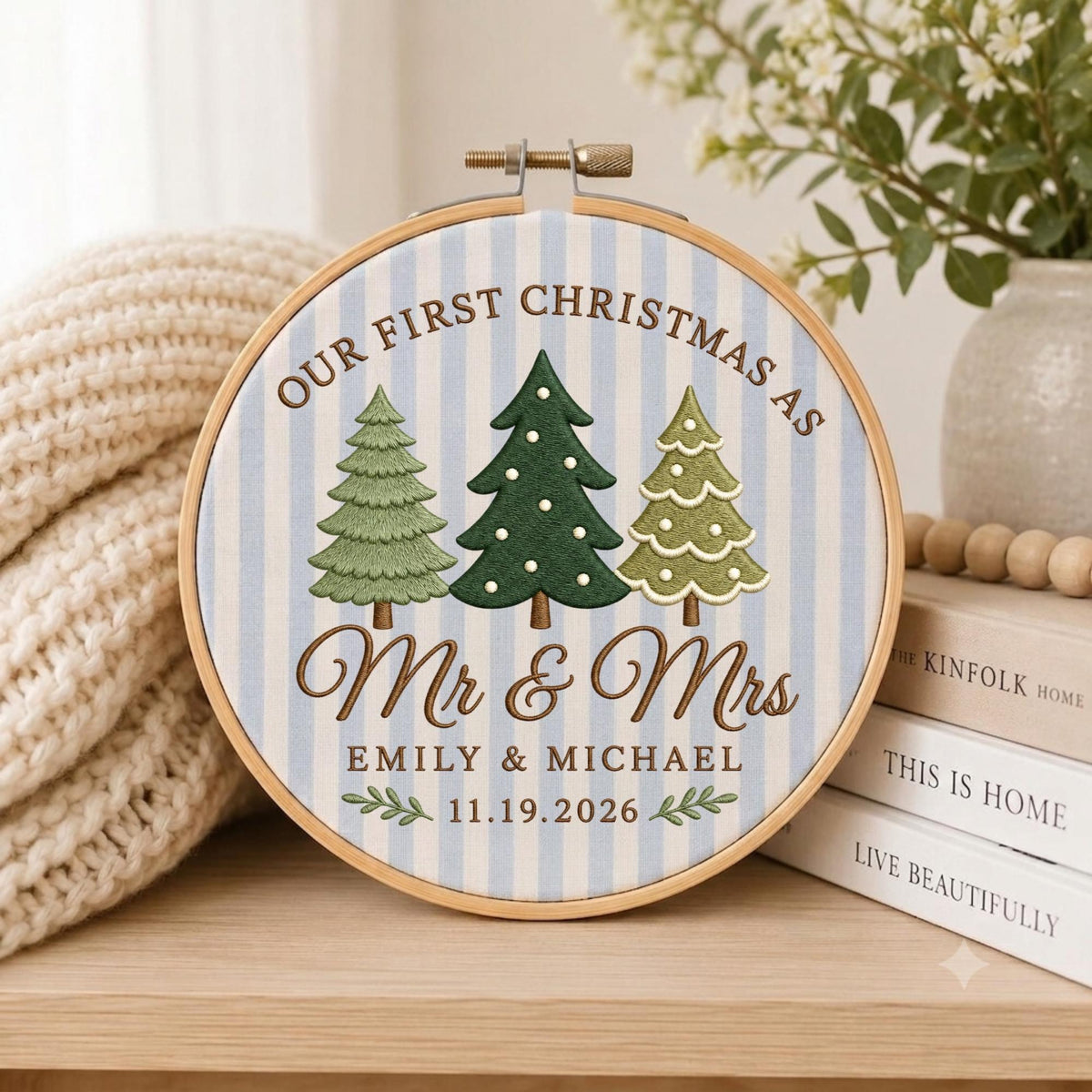 May include: A circular wooden embroidery hoop containing fabric with light blue and white vertical stripes. The embroidery features three green Christmas trees, the text "OUR FIRST CHRISTMAS AS Mr &amp; Mrs", "EMILY &amp; MICHAEL", and "11.19.2026".