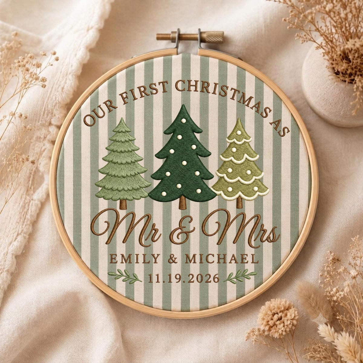 May include: A round wooden embroidery hoop containing fabric with vertical sage green and white stripes. The design features three embroidered Christmas trees, the text OUR FIRST CHRISTMAS AS Mr &amp; Mrs, the names EMILY &amp; MICHAEL, and the date 11.19.2026.