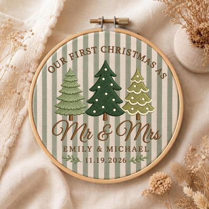 May include: A round wooden embroidery hoop containing fabric with vertical sage green and white stripes. The design features three embroidered Christmas trees, the text OUR FIRST CHRISTMAS AS Mr &amp; Mrs, the names EMILY &amp; MICHAEL, and the date 11.19.2026.