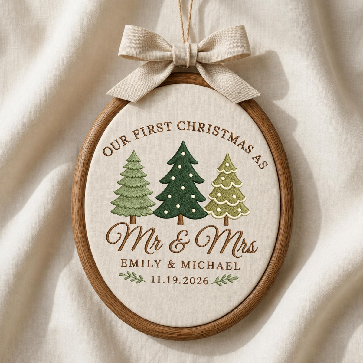May include: An oval wooden embroidery hoop containing off-white fabric with a beige ribbon bow at the top. The embroidery features three stylized Christmas trees, the text "OUR FIRST CHRISTMAS AS Mr &amp; Mrs", "EMILY &amp; MICHAEL", and "11.19.2026".