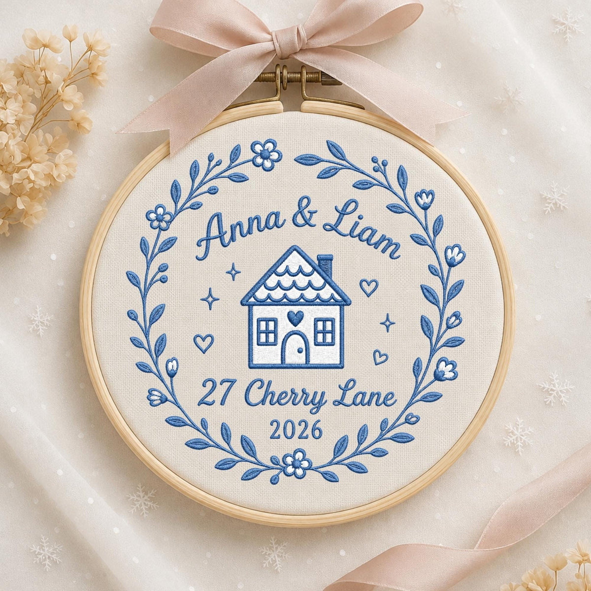 May include: A circular wooden embroidery hoop containing white fabric with blue thread embroidery. The design features a floral wreath surrounding a house icon, the text Anna &amp; Liam, 27 Cherry Lane, and 2026. A light pink ribbon is tied at the top.