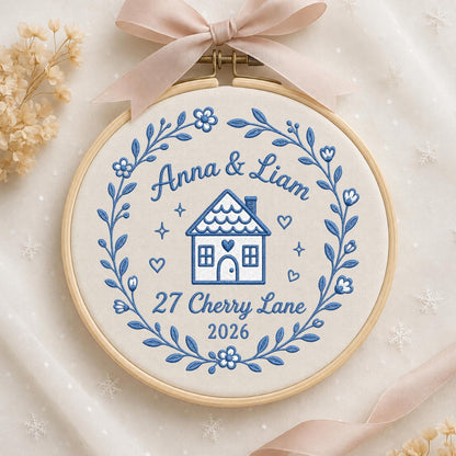May include: A circular wooden embroidery hoop containing white fabric with blue thread embroidery. The design features a floral wreath surrounding a house icon, the text Anna &amp; Liam, 27 Cherry Lane, and 2026. A light pink ribbon is tied at the top.
