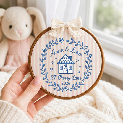 May include: A circular wooden embroidery hoop containing fabric with pink and white vertical stripes. The fabric features blue embroidery of a house, hearts, stars, and a floral wreath, with the text Anna &amp; Liam, 27 Cherry Lane, 2026.