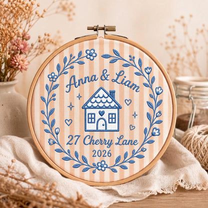 May include: A round wooden embroidery hoop containing fabric with vertical peach and white stripes. The fabric features blue embroidered text reading Anna &amp; Liam, 27 Cherry Lane, 2026, a central house icon, and a surrounding floral wreath.