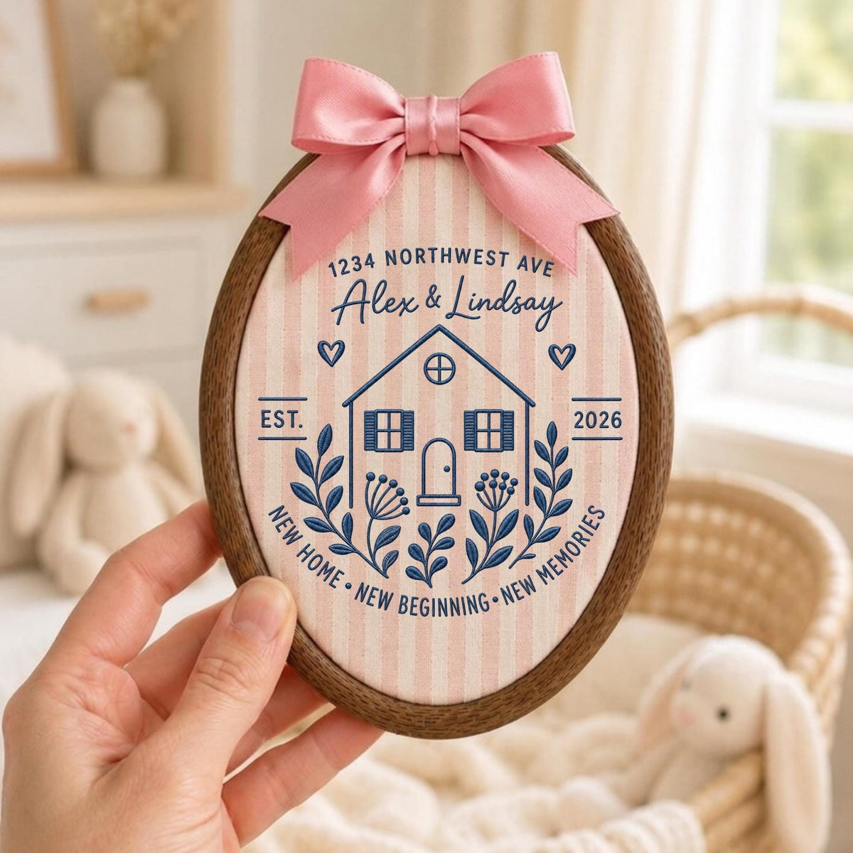 May include: An oval wooden embroidery hoop containing fabric with pink and white vertical stripes. The fabric features dark blue embroidery of a house, floral sprigs, hearts, and text reading 1234 NORTHWEST AVE, Alex &amp; Lindsay, EST. 2026, NEW HOME, NEW BEGINNING, NEW MEMORIES. A pink satin bow is attached to the top.