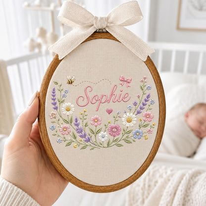 May include: An oval wooden embroidery hoop containing beige fabric with the name Sophie stitched in pink cursive. The design features embroidered pink and purple flowers, white daisies, green stems, a small bee, a pink heart, and a pink bow.