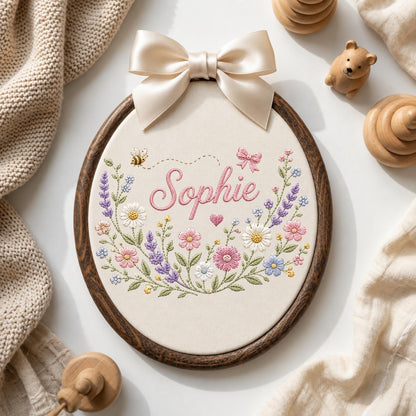 May include: An oval wooden embroidery hoop containing cream fabric with the name Sophie stitched in pink cursive. The design features colorful embroidered wildflowers, a small bee, a pink bow, and a heart. A cream satin bow is attached to the top.