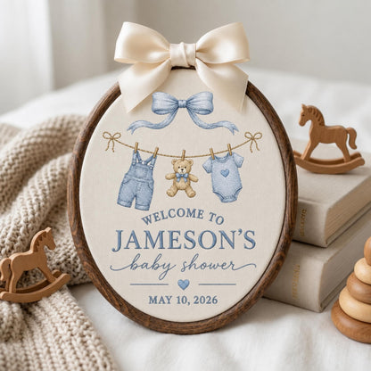 May include: An oval embroidery hoop sign with a dark wood frame and a cream fabric center. It features a light blue bow, a clothesline with denim overalls, a teddy bear, and a blue onesie. Text reads: WELCOME TO JAMESON'S baby shower MAY 10, 2026.