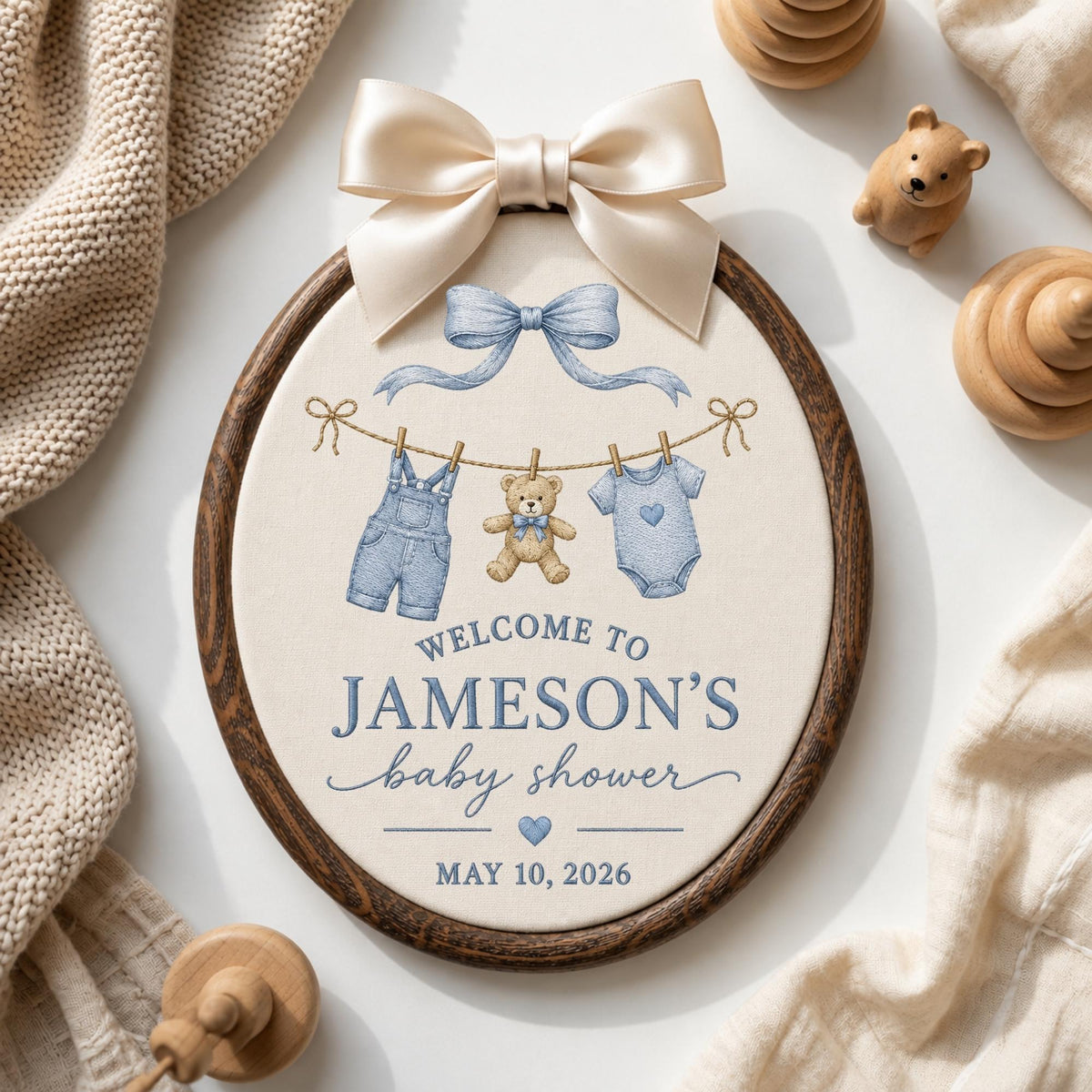 May include: An oval embroidery hoop wall hanging with a cream fabric center featuring a blue bow, a clothesline with denim overalls, a teddy bear, and a onesie. Text reads: WELCOME TO JAMESON'S baby shower MAY 10, 2026. Topped with a large satin bow.