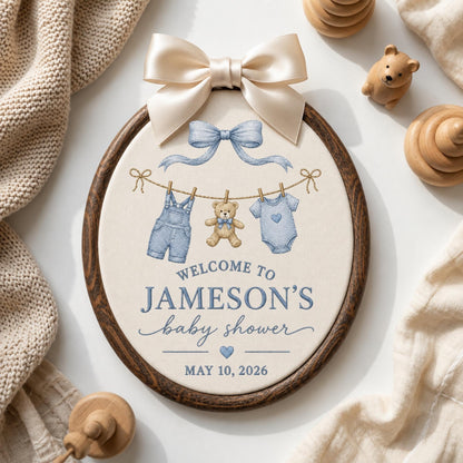 May include: An oval embroidery hoop wall hanging with a cream fabric center featuring a blue bow, a clothesline with denim overalls, a teddy bear, and a onesie. Text reads: WELCOME TO JAMESON'S baby shower MAY 10, 2026. Topped with a large satin bow.