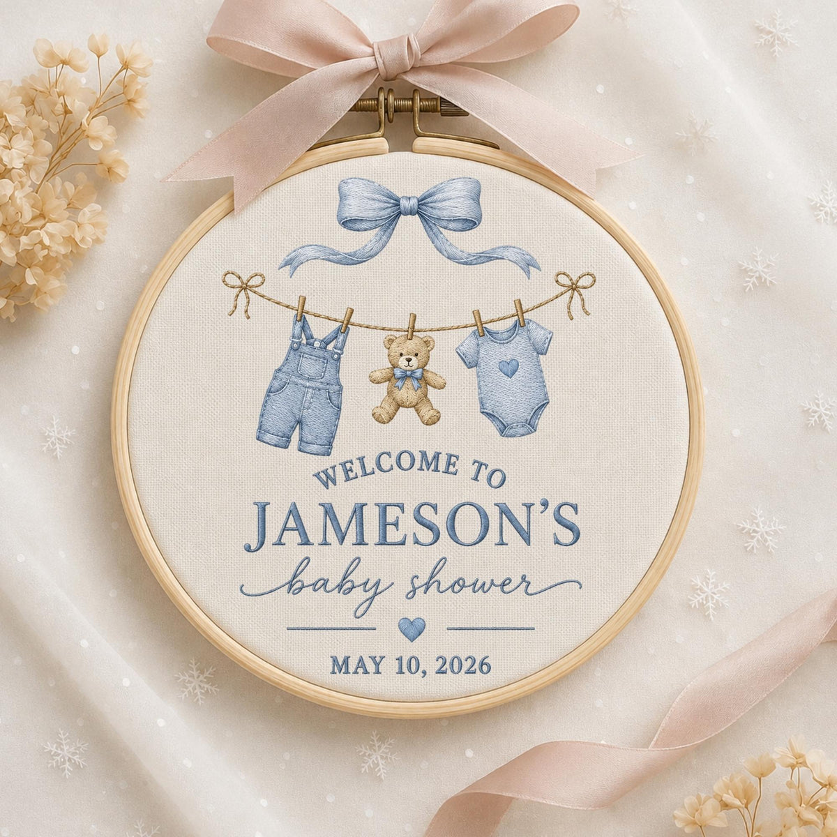 May include: A circular wooden embroidery hoop containing off-white fabric with a printed design of a blue bow, a teddy bear, and baby clothes on a clothesline. Text reads: WELCOME TO JAMESON'S baby shower MAY 10, 2026. A light pink ribbon is tied at the top.