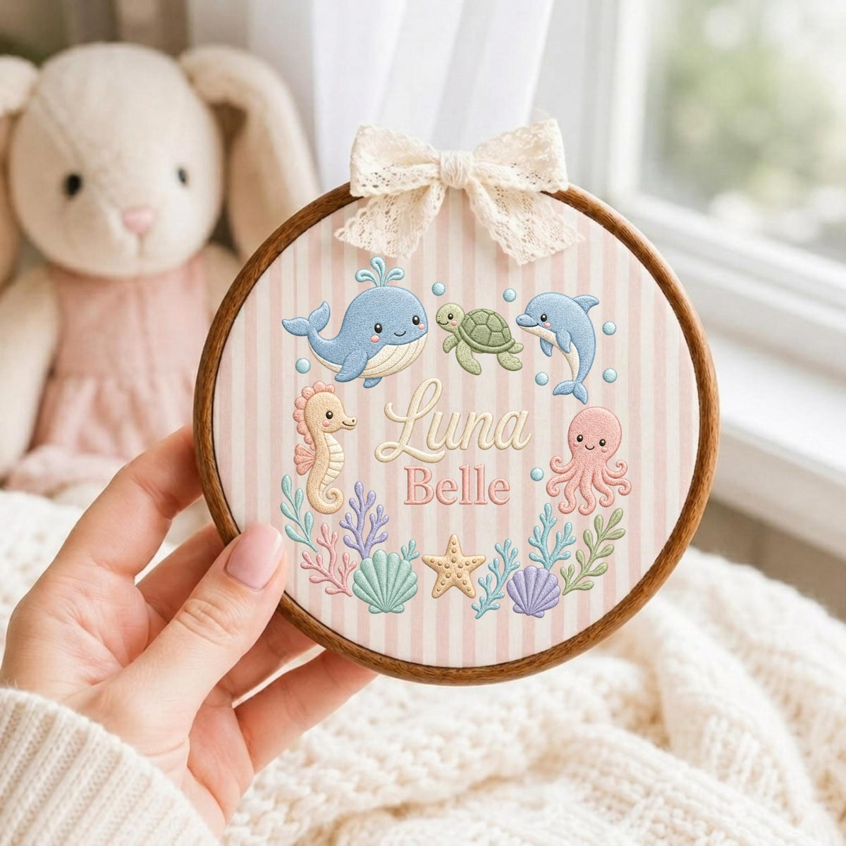 May include: A circular wooden embroidery hoop featuring a light pink and white striped fabric background. The design includes embroidered sea creatures: a blue whale, green turtle, blue dolphin, pink octopus, and seahorse, with the name Luna Belle.