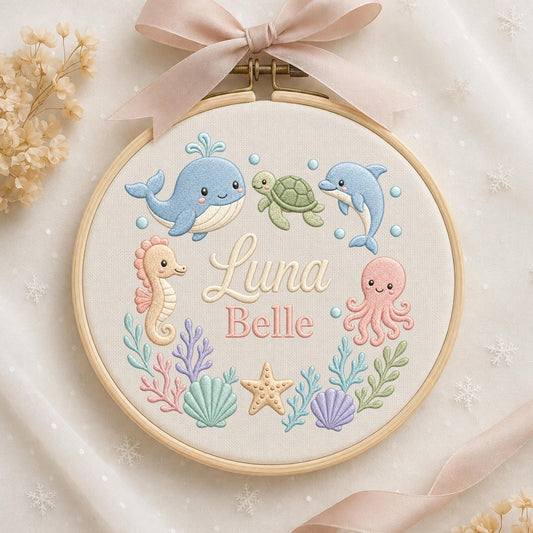 May include: A circular wooden embroidery hoop containing white fabric with a stitched design of a blue whale, green turtle, blue dolphin, peach seahorse, and pink octopus. The text Luna Belle is embroidered in the center with sea life accents.