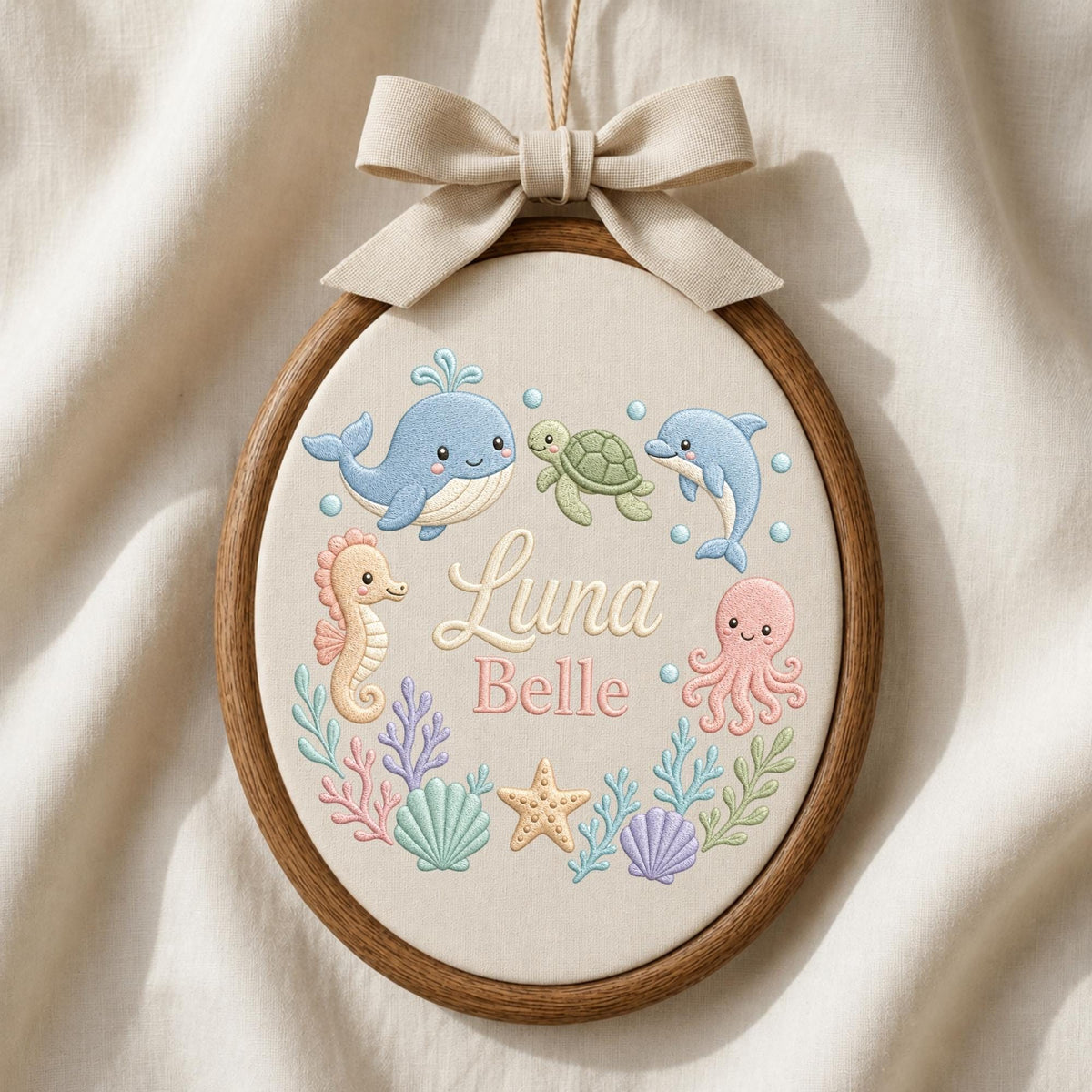 May include: An oval embroidery hoop featuring a beige fabric background with a light beige ribbon bow at the top. The design includes embroidered sea creatures: a blue whale, green turtle, blue dolphin, pink seahorse, and pink octopus, with the name Luna Belle in the center.