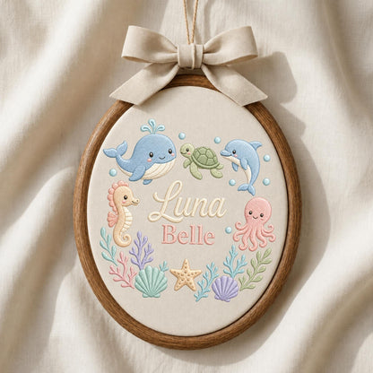 May include: An oval embroidery hoop featuring a beige fabric background with a light beige ribbon bow at the top. The design includes embroidered sea creatures: a blue whale, green turtle, blue dolphin, pink seahorse, and pink octopus, with the name Luna Belle in the center.