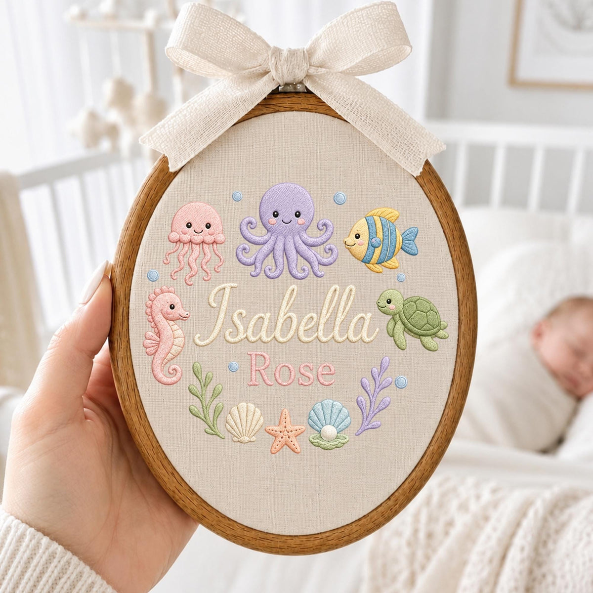 May include: An oval wooden embroidery hoop containing beige fabric with embroidered sea creatures including a pink jellyfish, purple octopus, yellow fish, green turtle, seahorse, shells, and starfish. The name Isabella Rose is stitched in the center.