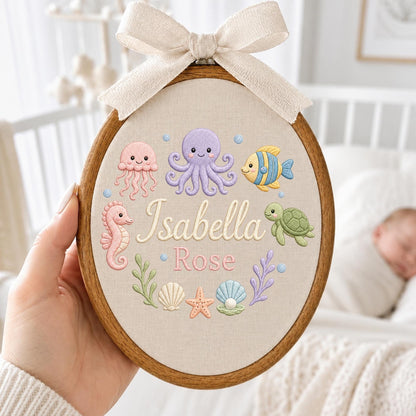 May include: An oval wooden embroidery hoop containing beige fabric with embroidered sea creatures including a pink jellyfish, purple octopus, yellow fish, green turtle, seahorse, shells, and starfish. The name Isabella Rose is stitched in the center.
