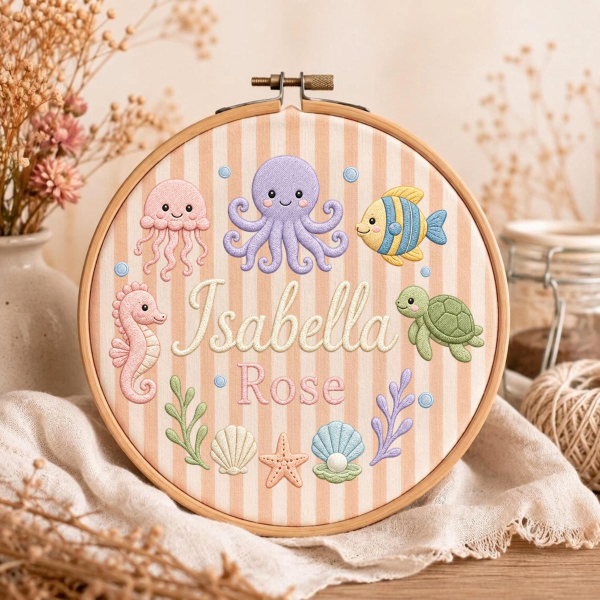 May include: A round wooden embroidery hoop containing fabric with vertical beige stripes. The fabric features embroidered sea creatures including a purple octopus, pink jellyfish, seahorse, green turtle, and fish, with the name Isabella Rose in white.
