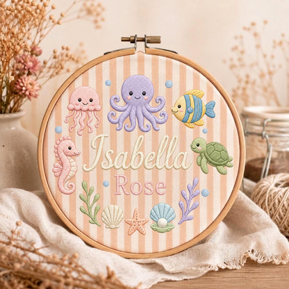 May include: A round wooden embroidery hoop containing fabric with vertical beige stripes. The fabric features embroidered sea creatures including a purple octopus, pink jellyfish, seahorse, green turtle, and fish, with the name Isabella Rose in white.