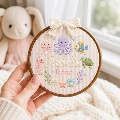 May include: A circular wooden embroidery hoop containing fabric with vertical pink and white stripes. The fabric features embroidered sea creatures including an octopus, jellyfish, seahorse, fish, and turtle, with the names Isabella Rose.