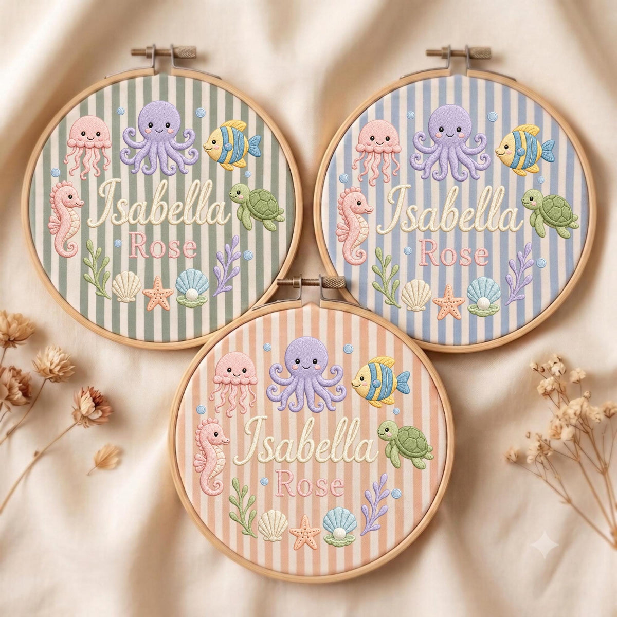 May include: Three circular wooden embroidery hoops featuring striped fabric backgrounds in sage green, light blue, and peach. Each hoop displays embroidered sea creatures including an octopus, jellyfish, seahorse, turtle, and shells with the name Isabella Rose.