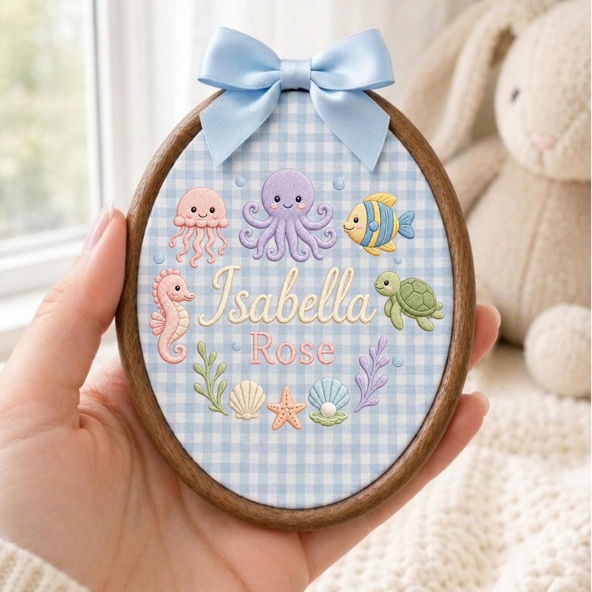May include: An oval wooden embroidery hoop featuring a light blue gingham fabric background. The design includes embroidered sea creatures like a purple octopus, pink jellyfish, yellow fish, and green turtle, with the name Isabella Rose in script.