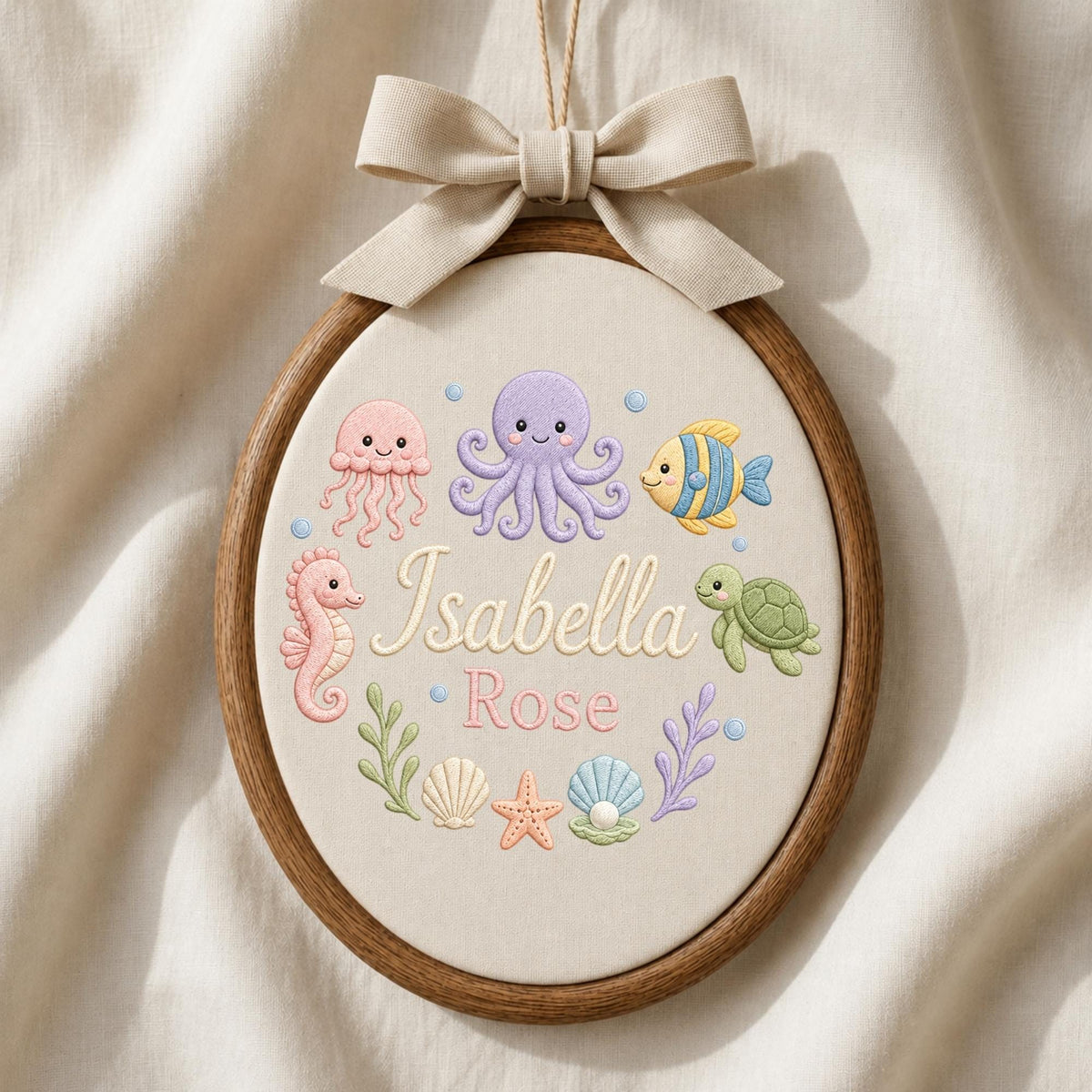 May include: An oval wooden embroidery hoop containing fabric with stitched sea creatures including a pink jellyfish, purple octopus, yellow fish, and green turtle. The text Isabella Rose is embroidered in the center with seashells and seaweed.