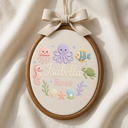 May include: An oval wooden embroidery hoop containing fabric with stitched sea creatures including a pink jellyfish, purple octopus, yellow fish, and green turtle. The text Isabella Rose is embroidered in the center with seashells and seaweed.
