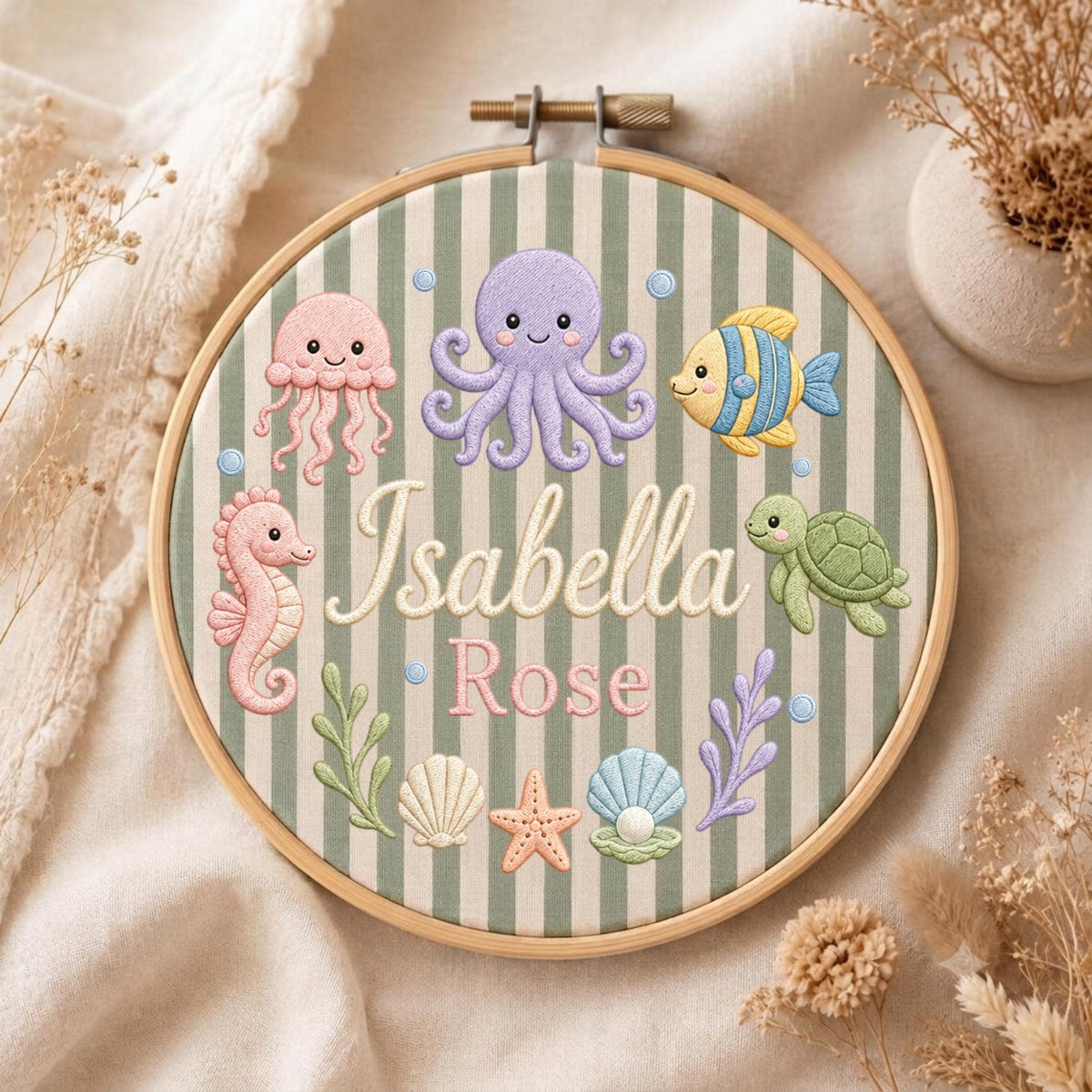 May include: A round wooden embroidery hoop containing fabric with vertical sage green and white stripes. The fabric features embroidered sea creatures including a purple octopus, pink jellyfish, yellow fish, green turtle, seahorse, starfish, and shells. The name Isabella Rose is embroidered in the center.