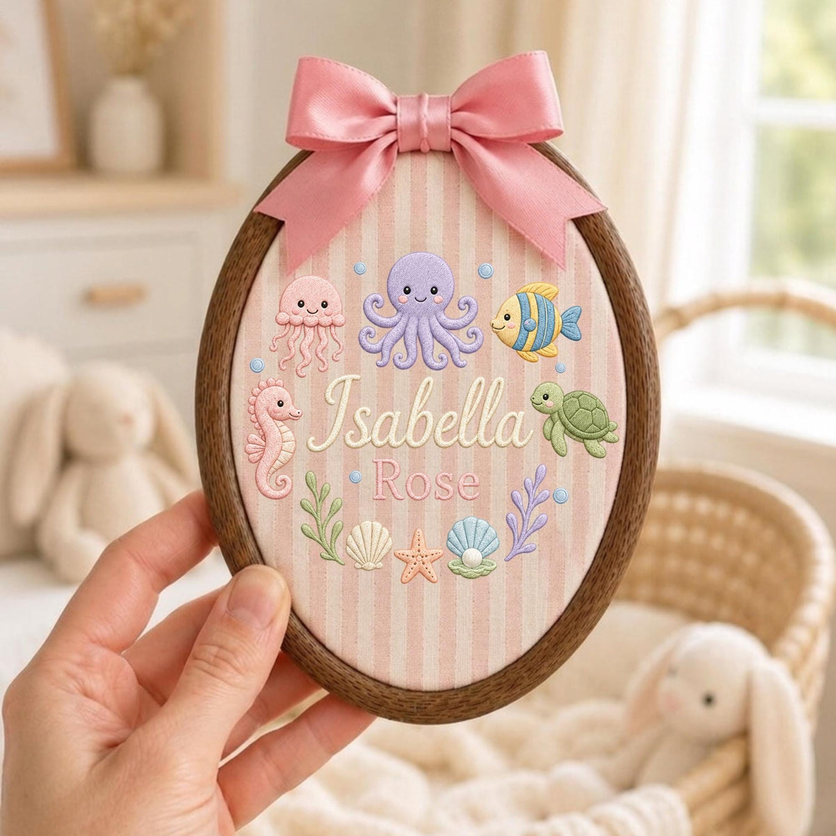 May include: An oval wooden embroidery hoop containing fabric with vertical pink stripes. The fabric features embroidered sea creatures, shells, and the name Isabella Rose in white script. A pink satin ribbon bow is attached to the top.