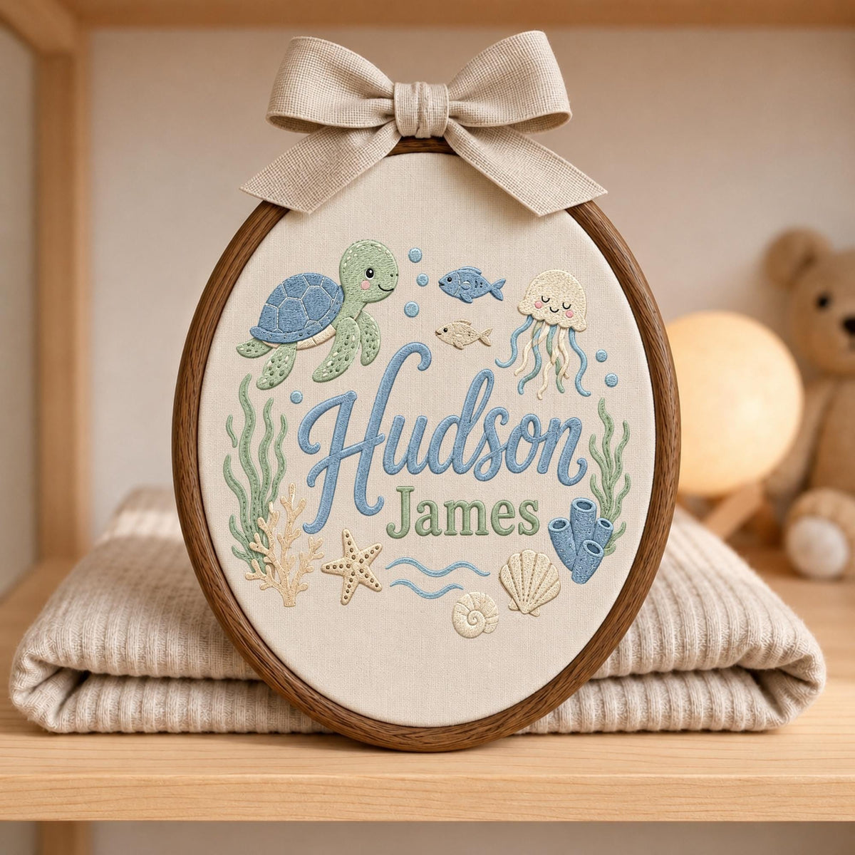 May include: An oval embroidery hoop featuring a beige fabric background with the embroidered name Hudson James in blue and green script. The design includes a green sea turtle, a jellyfish, fish, coral, a starfish, and seashells in soft pastel tones.