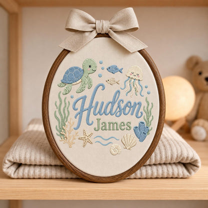 May include: An oval embroidery hoop featuring a beige fabric background with the embroidered name Hudson James in blue and green script. The design includes a green sea turtle, a jellyfish, fish, coral, a starfish, and seashells in soft pastel tones.