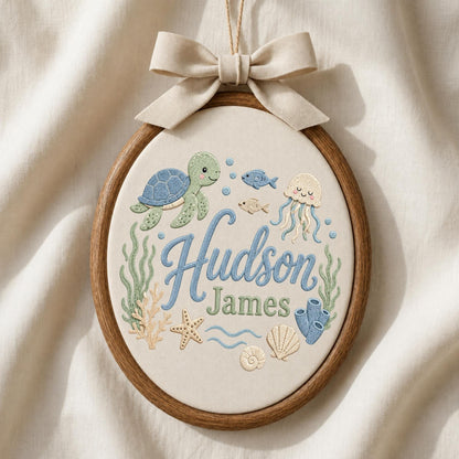 May include: An oval wooden embroidery hoop containing cream fabric with the embroidered names Hudson James in blue and green. The design features a green sea turtle, a jellyfish, a starfish, coral, seashells, and small fish in blue and tan.