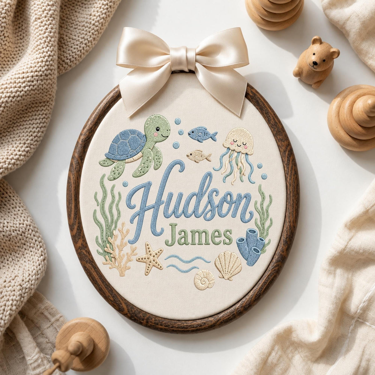 May include: An oval embroidery hoop featuring a dark wood frame and a cream fabric center. The embroidery depicts a sea turtle, jellyfish, fish, starfish, and shells in blue, green, and tan. The name Hudson James is stitched in blue and green.