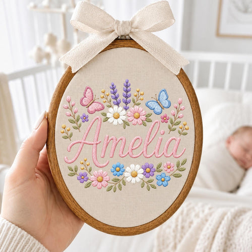 May include: An oval wooden embroidery hoop containing a beige fabric panel with the name Amelia stitched in pink cursive script. The design features embroidered colorful flowers, green leaves, and two butterflies, topped with a beige fabric bow.