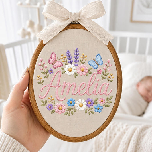 May include: An oval wooden embroidery hoop containing a beige fabric panel with the name Amelia stitched in pink cursive script. The design features embroidered colorful flowers, green leaves, and two butterflies, topped with a beige fabric bow.