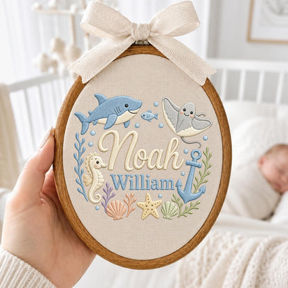 May include: An oval wooden embroidery hoop containing fabric with embroidered sea creatures including a shark, stingray, seahorse, starfish, and anchor. The names Noah and William are stitched in the center, topped with a beige fabric bow.