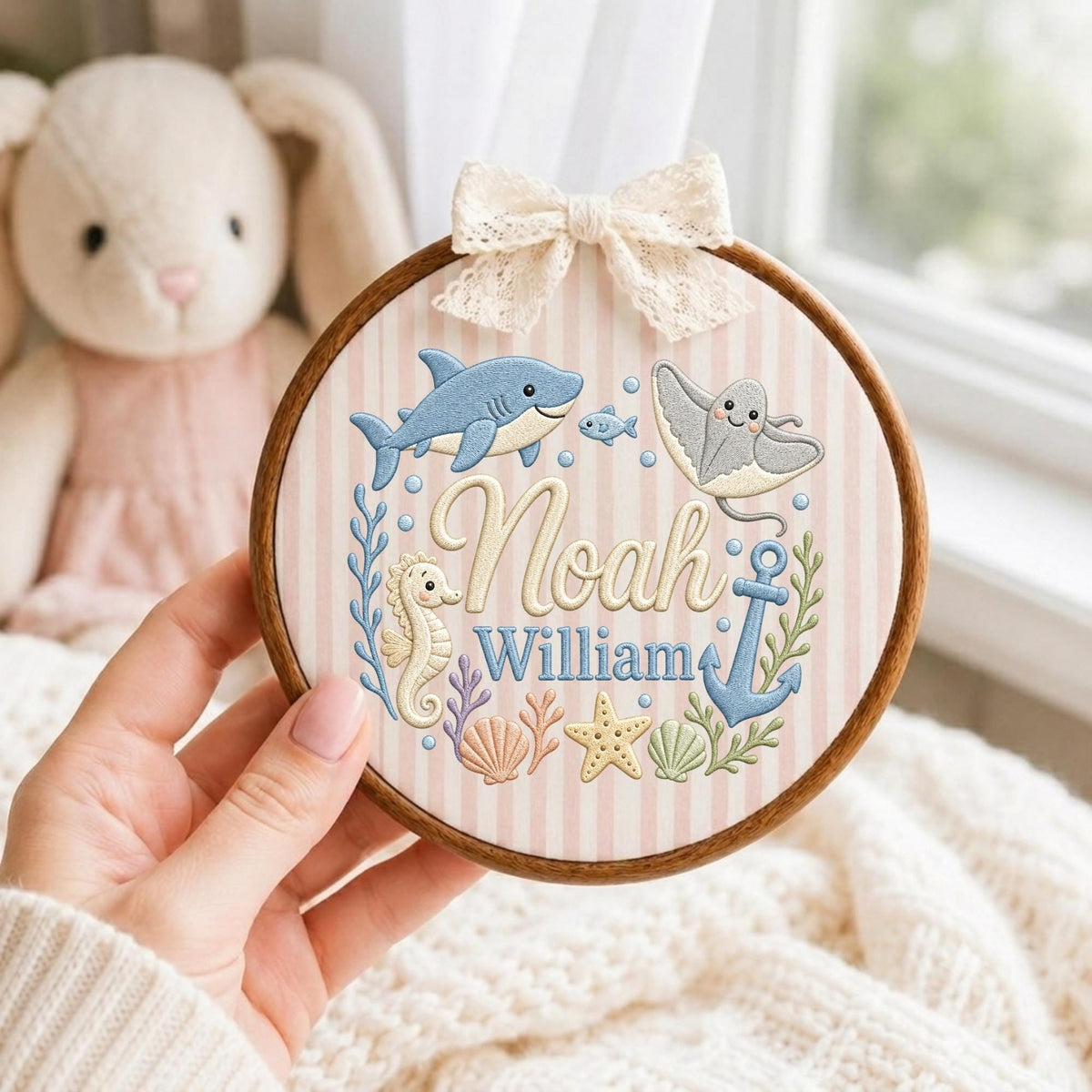 May include: A circular wooden embroidery hoop featuring a light pink and white striped fabric background. The design includes embroidered sea creatures, a blue anchor, and the names Noah William in script. A cream lace bow sits at the top.
