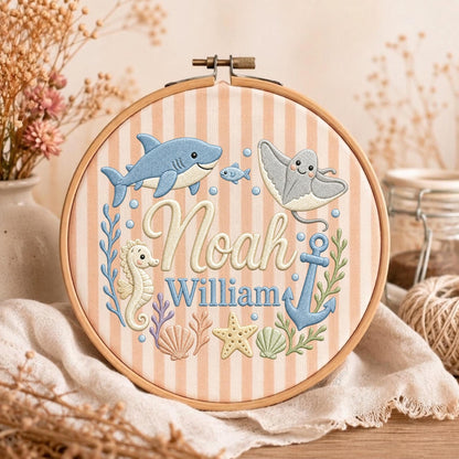 May include: A round wooden embroidery hoop containing fabric with vertical peach and white stripes. The fabric features embroidered sea creatures including a blue shark, a stingray, a seahorse, a starfish, and an anchor, with the name Noah William.