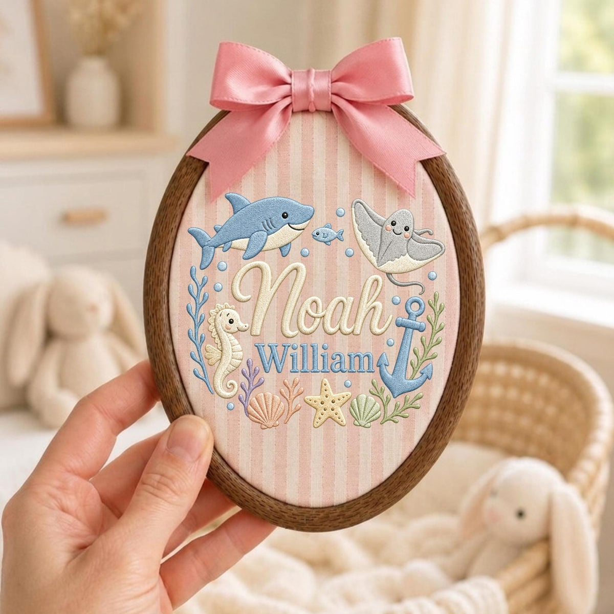 May include: An oval wooden embroidery hoop containing fabric with pink and white vertical stripes. The fabric features embroidered sea creatures, an anchor, and the names Noah and William. A pink satin ribbon bow is attached to the top.