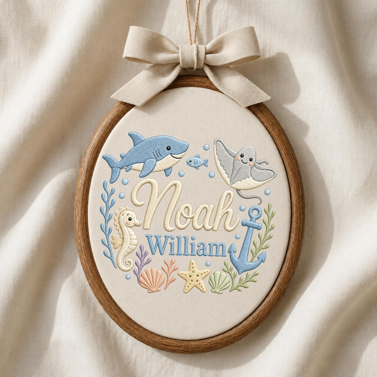 May include: An oval wooden embroidery hoop containing cream fabric with stitched sea life including a blue shark, a stingray, a seahorse, a starfish, a seashell, and an anchor. The names Noah and William are embroidered in the center with a beige bow.