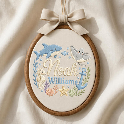 May include: An oval wooden embroidery hoop containing cream fabric with stitched sea life including a blue shark, a stingray, a seahorse, a starfish, a seashell, and an anchor. The names Noah and William are embroidered in the center with a beige bow.