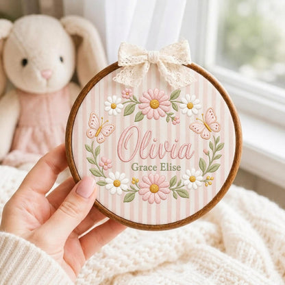 May include: A circular wooden embroidery hoop featuring a light pink and white striped fabric background. The center displays the embroidered name Olivia Grace Elise in pink script and grey print, surrounded by pink daisies, green leaves, and butterflies.