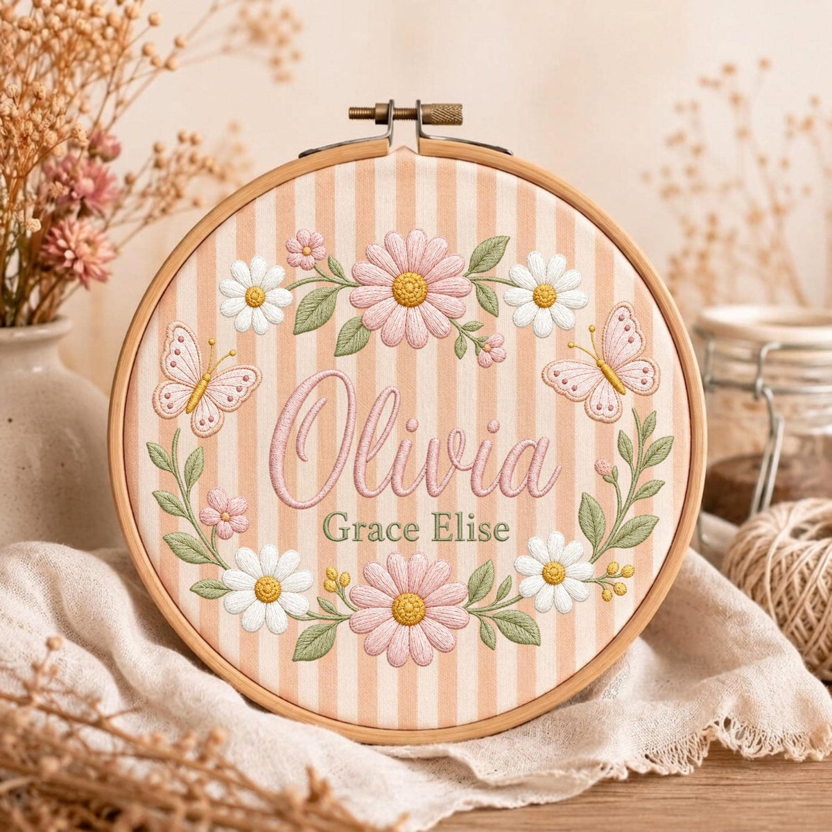 May include: A circular wooden embroidery hoop containing fabric with vertical peach and white stripes. The fabric features embroidered pink and white daisies, green leaves, and two pink butterflies. The name Olivia Grace Elise is stitched in pink.