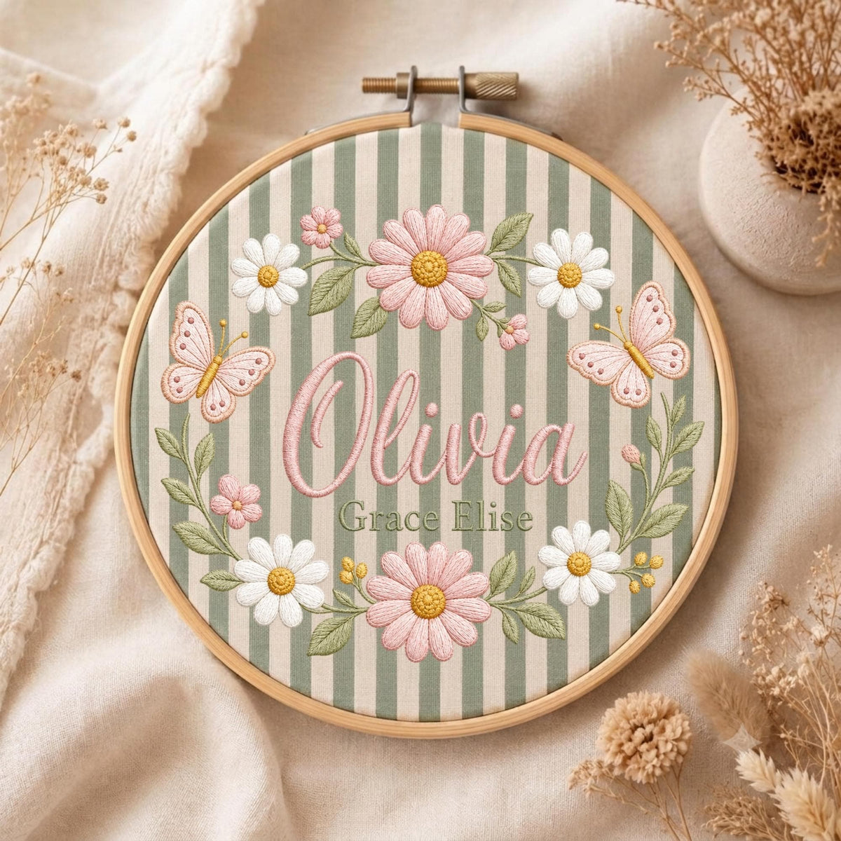 May include: A circular wooden embroidery hoop containing fabric with vertical sage green and white stripes. The embroidery features the name Olivia in pink script, the name Grace Elise in green text, pink daisies, white daisies, and two pink butterflies.